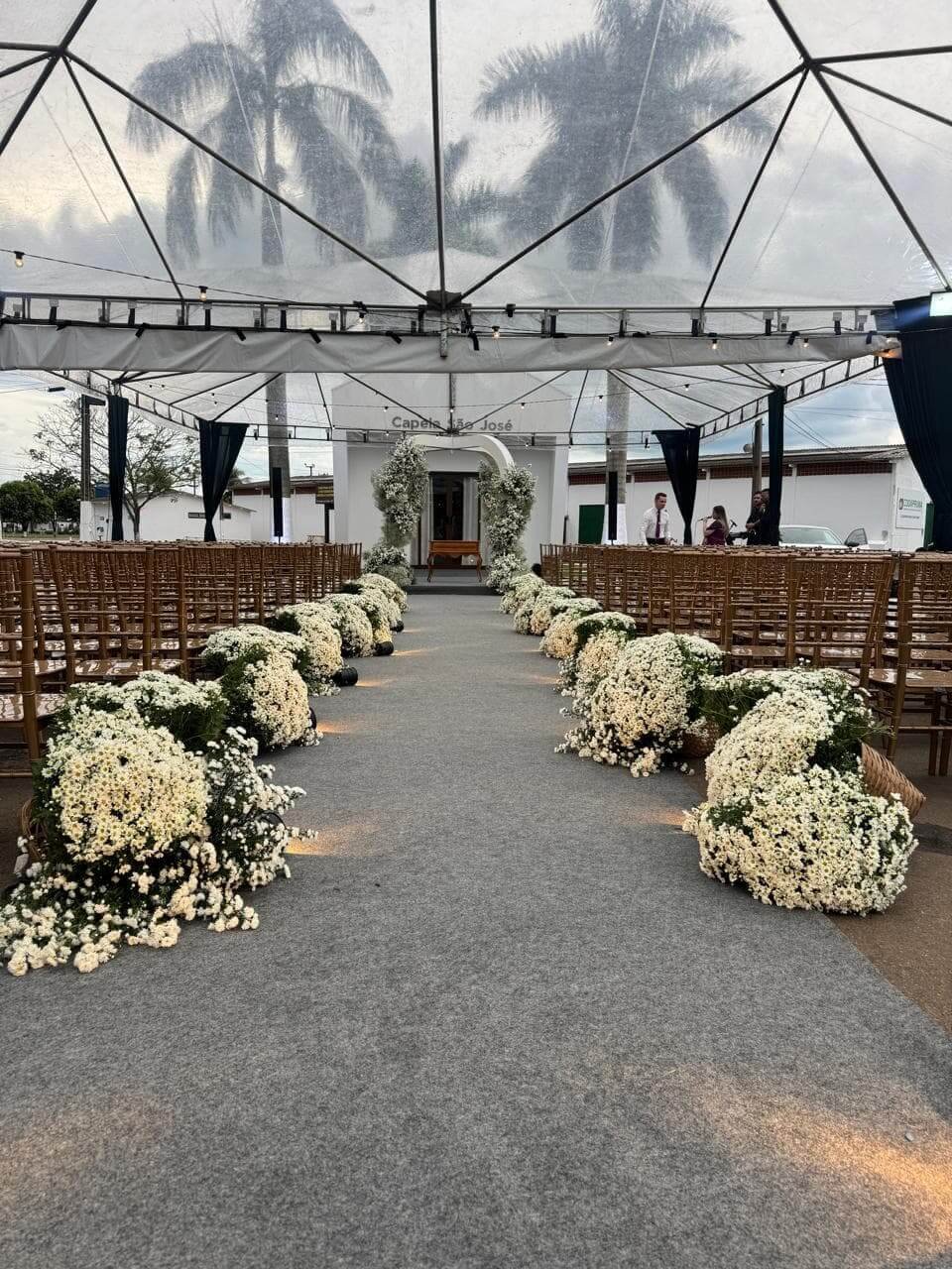 Cerimônia de casamento ao ar livre com estrutura de cobertura transparente (tenda), cadeiras de madeira e corredor de flores brancas.