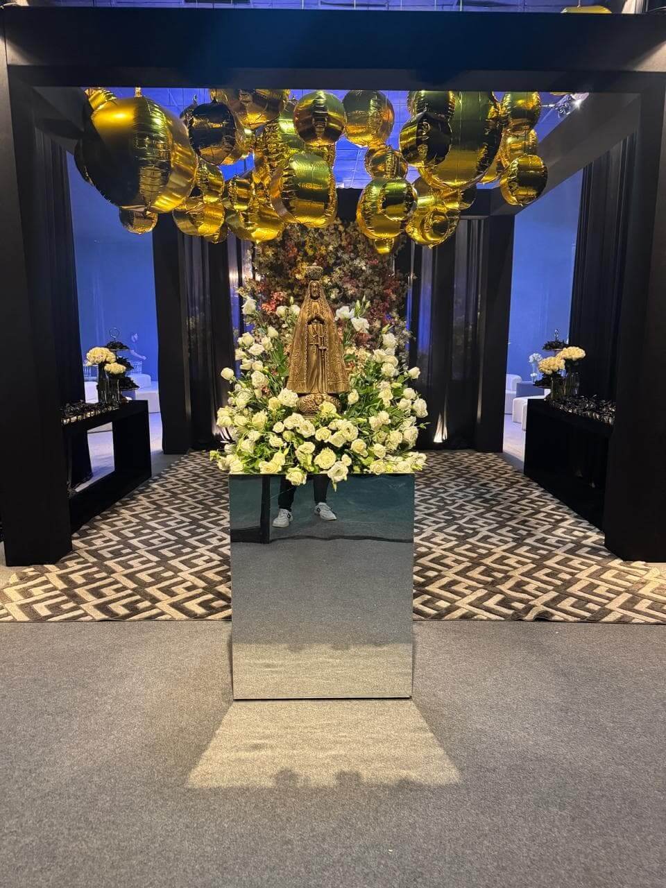 spaço decorado com tema religioso, apresentando imagem de Santa em pedestal espelhado, flores brancas e balões dourados no teto.