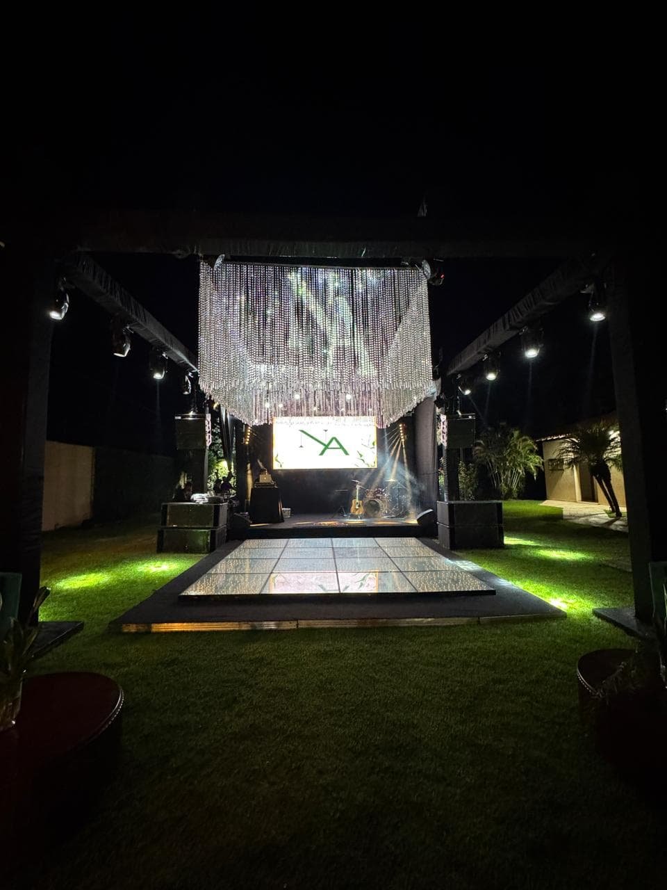 Área de cerimônia noturna ao ar livre com passarela espelhada, cadeiras brancas, lustre de cristal e iluminação cênica dramática.