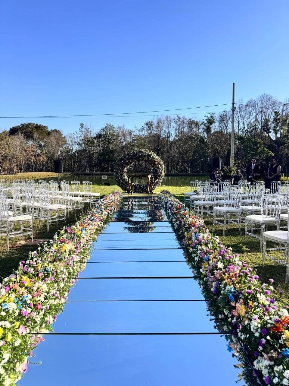 Cerimônia de casamento diurna ao ar livre com longa passarela espelhada, arranjos florais coloridos nas laterais e arco floral ao fundo.