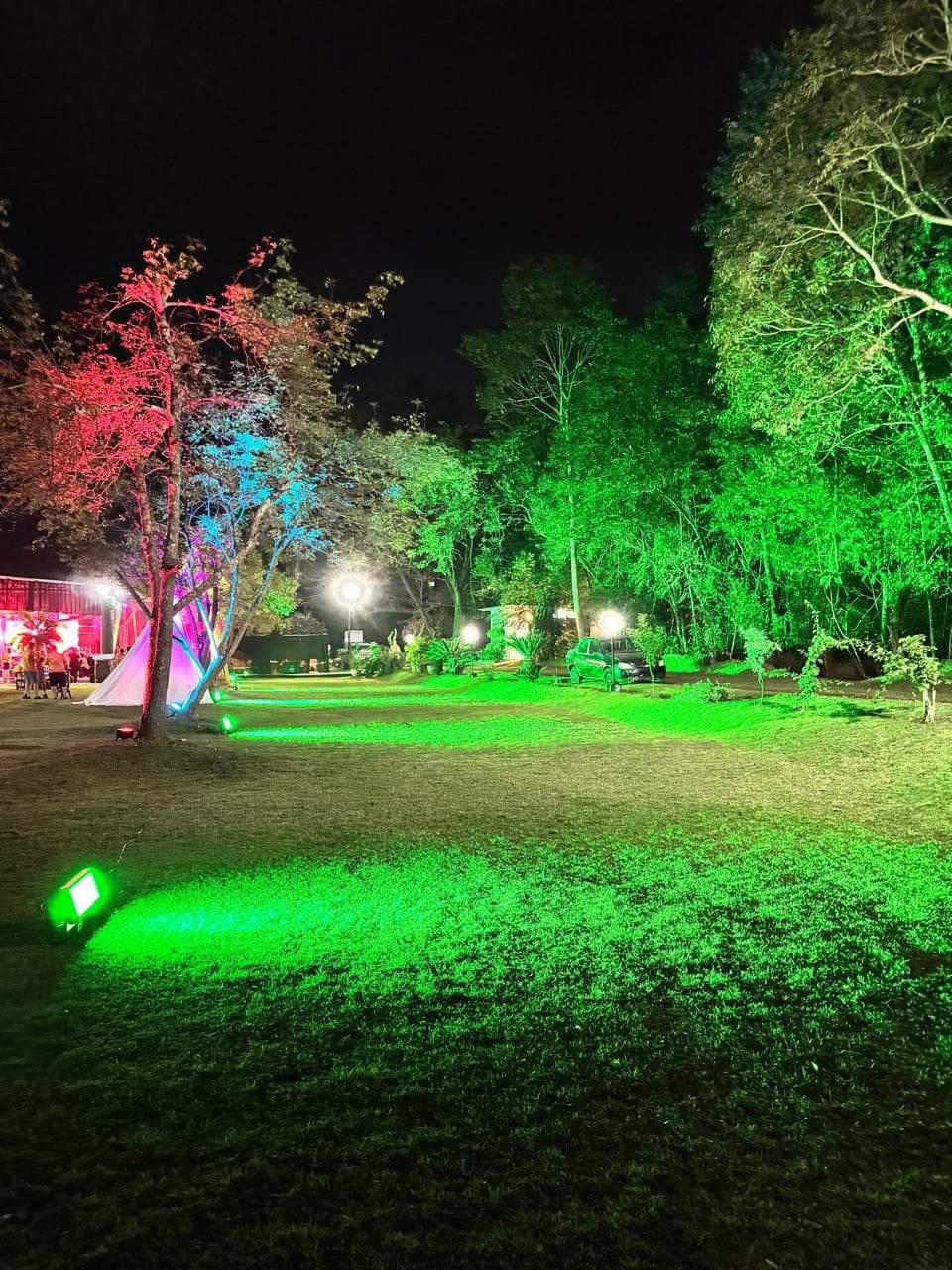 Iluminação cênica externa noturna destacando árvores e paisagismo com luzes verdes e vermelhas, criando um ambiente mágico.