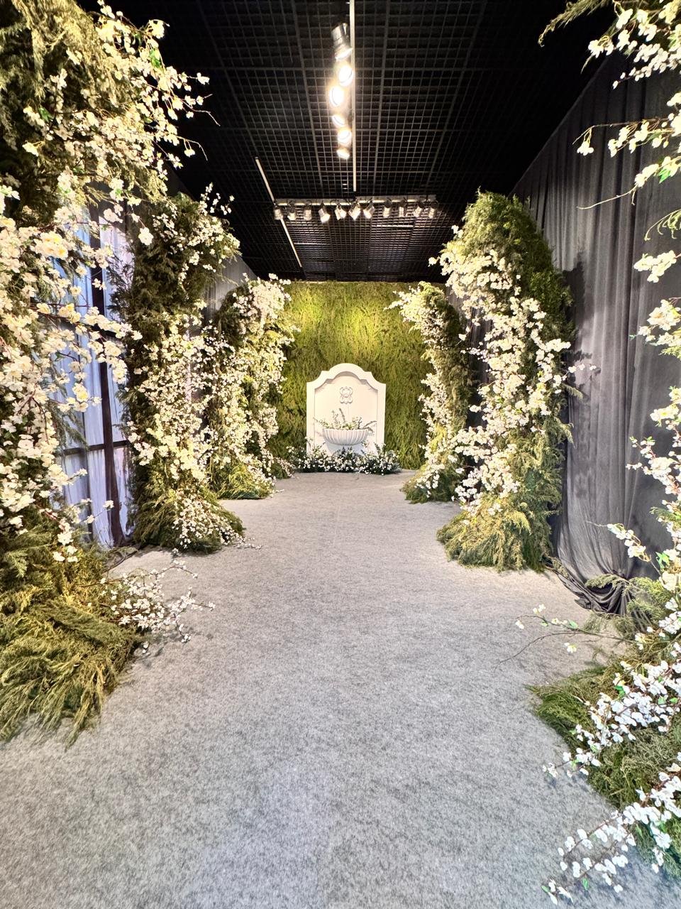 Corredor de cerimônia interna decorado com paredes verdes (muro inglês), flores brancas e iluminação pontual.