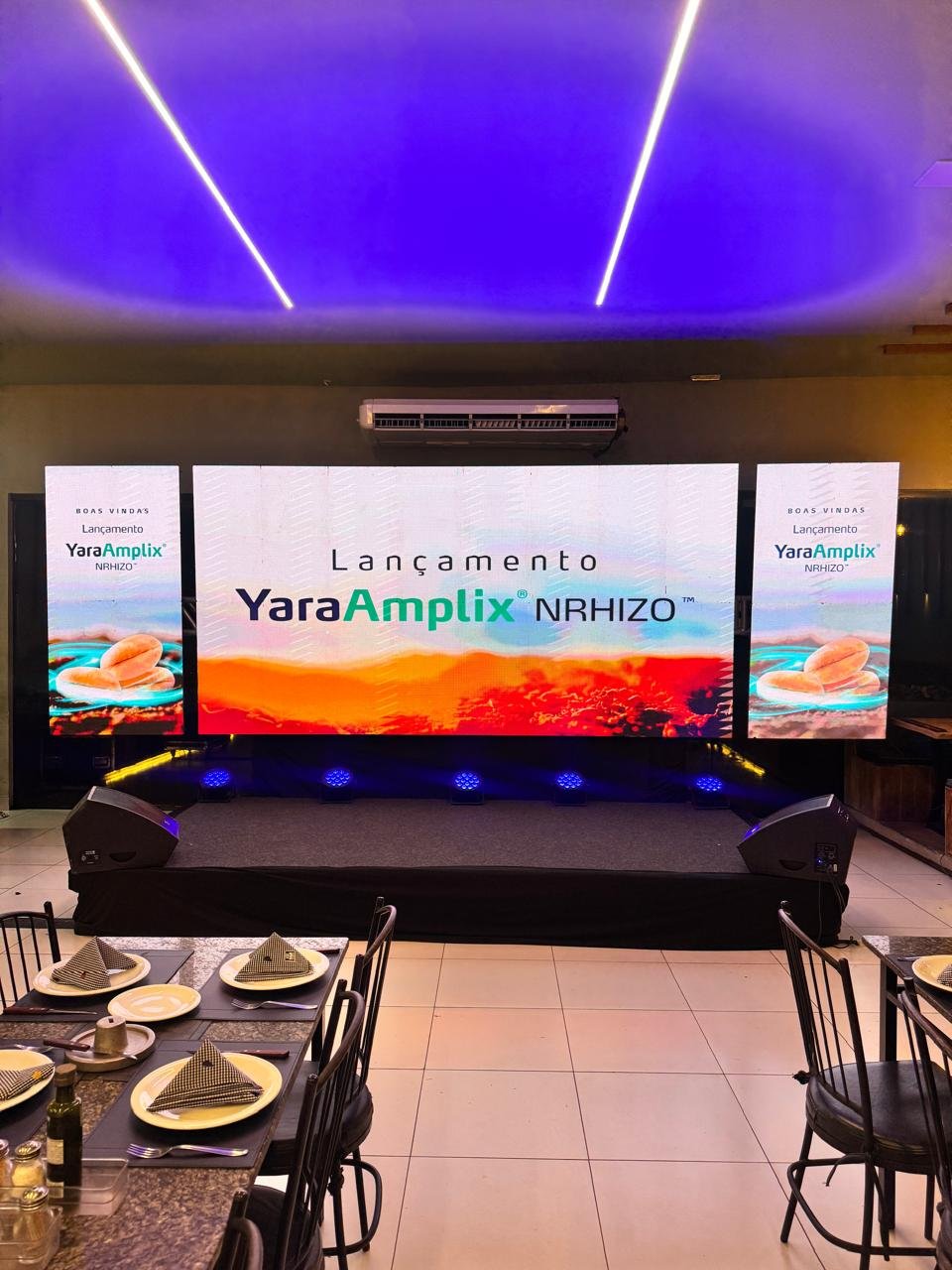 Palco corporativo com painel de LED triplo exibindo apresentação de lançamento de produto (YaraAmplix).