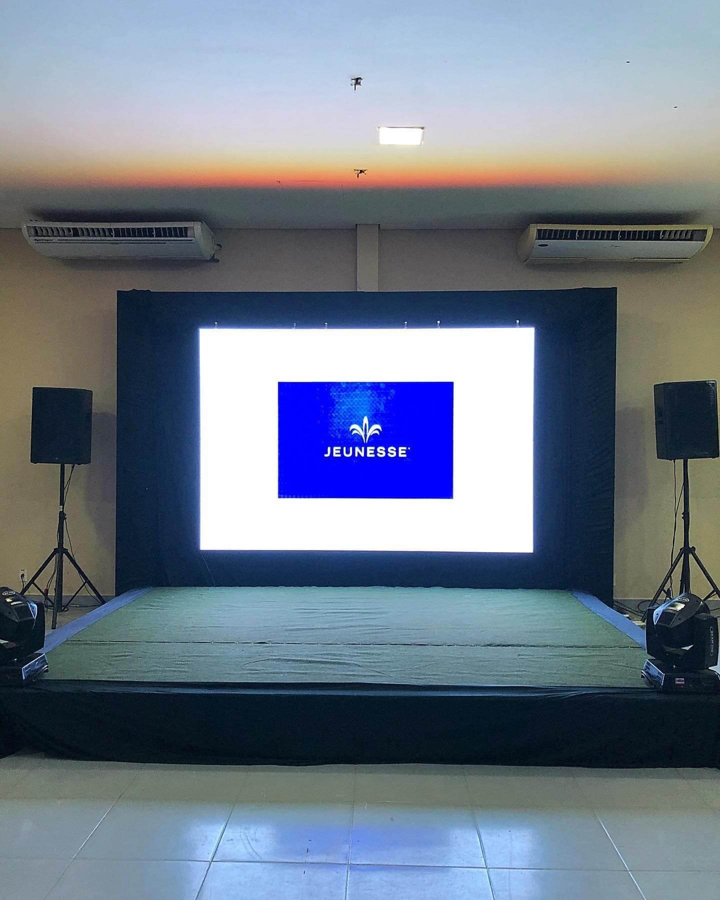 Palco para convenções e palestras com carpete verde, grande painel de LED central de alta definição (exibindo logo Jeunesse) e sistema de som.